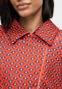 Κόκκινο παλτό με σχέδιο, διαθέτει μεγάλο γιακά, διαμάντι σχήματα σε μωβ και λευκό, και μπροστινό φερμουάρ. Το ύφασμα έχει λεία υφή.