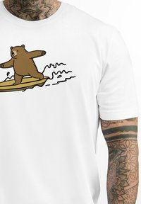 White t-shirt featuring a cartoon bear surfing on a yellow board, with black wave lines around it. Textured, short-sleeved material.