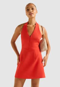 Crvena halter haljina s dubokim V-izrezom, pripijenim korzetom i raširenom suknjom. Model drži zlatnu torbicu s teksturom. Gladak materijal, minimalni detalji.