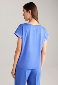Blauwe T-shirt met korte mouwen, in een ontspannen pasvorm, met een gladde textuur en een subtiele logoprint op de achterkant. Gecombineerd met blauwe broeken.