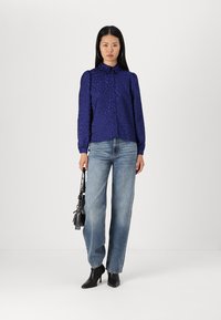 Asymmetric button-up shirt in deep purple with a textured heart pattern, paired with high-waisted light blue jeans and black heeled boots.