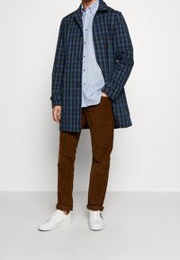 Cappotto impermeabile a quadretti blu e neri con chiusura a bottoni, indossato sopra una camicia azzurra chiara e pantaloni marroni, completato da sneakers bianche.