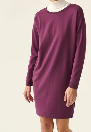 Frau trägt ein langärmeliges Kleid in Pflaumenfarbe über einem weißen Rollkragen, steht vor einem einfarbigen hellen Hintergrund.