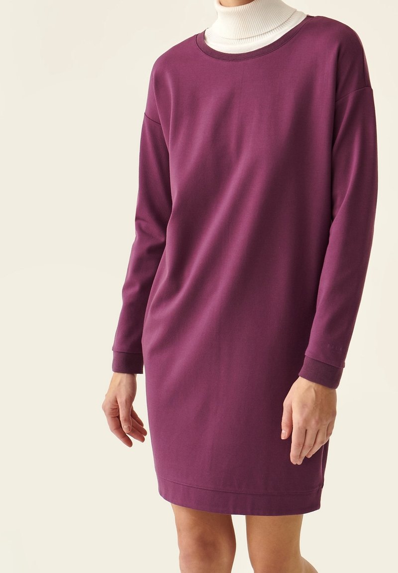 Femme portant une robe prune à manches longues par-dessus un col roulé blanc, debout devant un fond clair uni.