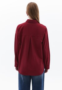 Rotes Langarmhemd mit Kragen, mit einer Faltenstruktur am Rücken und knöpfbaren Manschetten, kombiniert mit lockeren blauen Jeans.