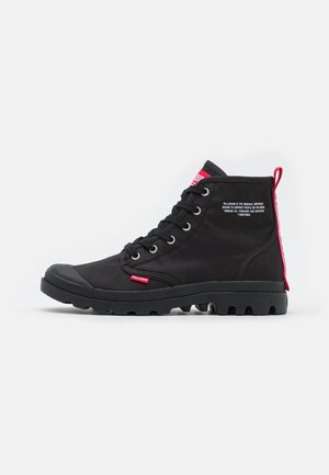 Bottes de cheville en toile noire avec capuchon en caoutchouc, lacets à l'avant et semelle texturée. Présente une étiquette de tirage rouge bien visible et un détail de logo.