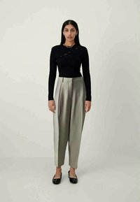Black long-sleeve top with diagonal detailing, paired with high-waisted, light grey pleated pants and black flat shoes. Minimalistic design.
