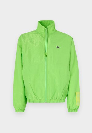 Veste coupe-vent verte avec un col montant, fermeture éclair à l'avant, poignets élastiques et ourlet. Présente un petit logo et un accent en étiquette sur la manche.