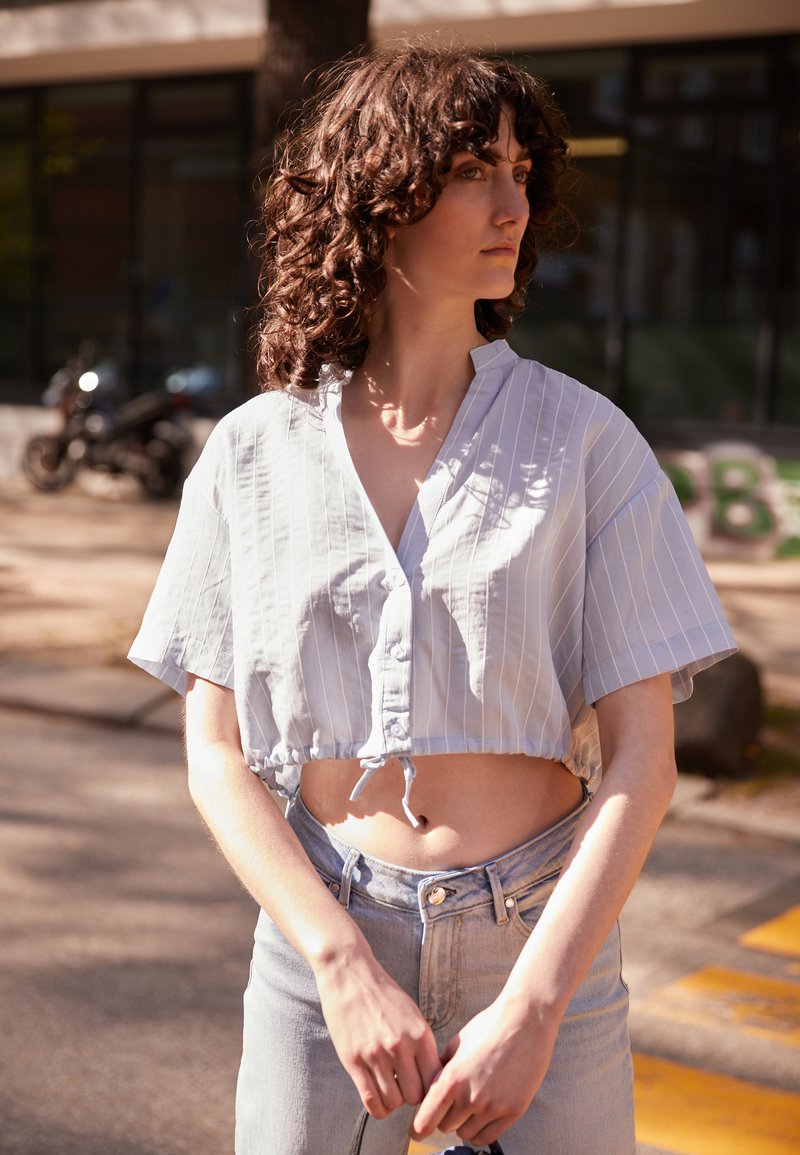 Blusa curta riscada em azul claro com mangas curtas e fecho de botões, combinada com jeans de cintura alta em denim claro. Modelo em pé ao ar livre.