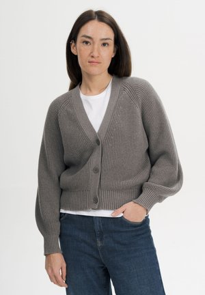 Woman with long dark hair wearing a gray knitted buttoned cardigan over a white shirt and blue jeans, standing with one hand in pocket.