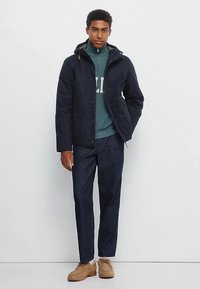 Marineblå vandtæt jakke med hætte, forlommer og lynlås. Kombineret med en grøn sweater, mørke jeans og brune sko.