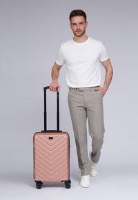 Hombre con camiseta blanca y pantalones grises caminando con una maleta de ruedas en color oro rosa, sosteniendo el mango extendido.
