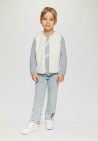 Gilet in pelliccia sintetica bianca sopra una camicia a maniche lunghe azzurra chiara con un motivo a cuore, abbinato a jeans azzurri chiari e scarpe da ginnastica bianche.