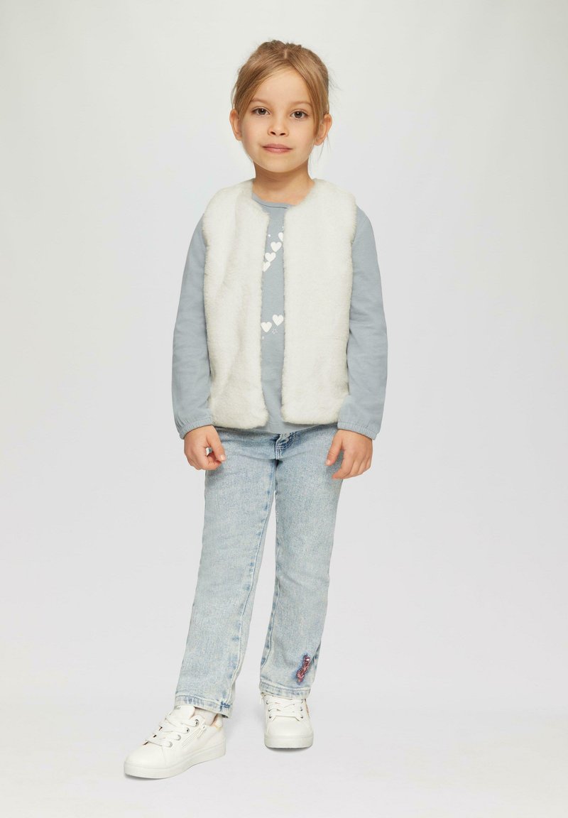 Gilet in pelliccia sintetica bianca sopra una camicia a maniche lunghe azzurra chiara con un motivo a cuore, abbinato a jeans azzurri chiari e scarpe da ginnastica bianche.