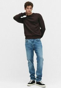 Bruine gebreide sweater, lichtblauwe jeans, zwart-witte sneakers. Model staat met één hand op de nek en toont een casual outfit en fit.