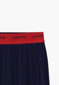Námořnická plisovaná sukně s červeným elastickým pasem s nápisem "LACOSTE". Hladká textura a jednotné plisování vytváří strukturovaný vzhled.