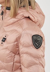 Donna che indossa una giacca imbottita a sacco rosa con cappuccio e una toppa nera a forma di scudo sulla manica.
