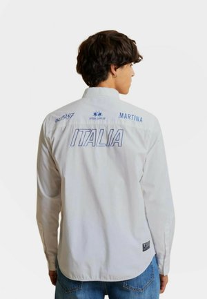 Jonge man met kort donker haar draagt een wit shirt met lange mouwen met de tekst "Italia," "Martina" en "Est. 1987" op de rug, gecombineerd met blauwe jeans.