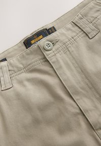 Shorts en coton beige avec fermeture à bouton, passants pour ceinture et étiquette "Next Durable Goods". Texture douce et coupe droite.