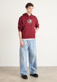 Felpa con cappuccio bordeaux con un logo multicolore e lacci, abbinata a jeans a gamba larga blu chiaro e scarpe nere senza lacci.