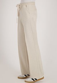 Beige wijde broek met elastische tailleband en trekkoord, gedragen met witte sneakers met zwarte strepen en rubberen zolen.