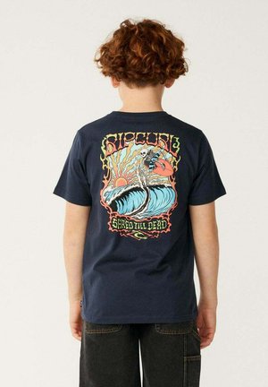 Niño con cabello rizado que lleva una camiseta azul marino con un dibujo colorido de un esqueleto surfeando una ola y el texto "Shred Till Dead" en la espalda.