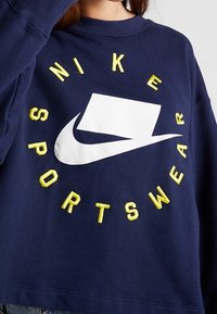 Marinblå sweatshirt med en rund vit Nike-logotyp och gul broderad text "SPORTSWEAR". Har en rund halsringning och avslappnad passform.
