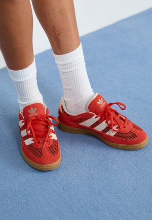 Red sneaker with white stripes, made of mesh and suede, featuring a rubber sole. Worn with white ribbed socks. Background is blue.