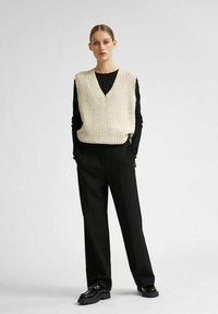 Pull sans manches en tricot écru avec un décolleté en V, porté par-dessus un haut noir à manches longues. Assorti à un pantalon noir ajusté et des chaussures.