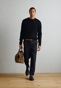 Pulover negru cu dungi, pantaloni negri, centură maronie, ținând o geantă din pânză maronie, purtând pantofi negri, stând pe un podea din lemn cu un fundal deschis.