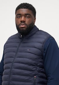 Gilet rembourré bleu marine avec coutures verticales, col montant et fermeture éclair devant. Comprend un petit logo près de la poitrine et des poches latérales.