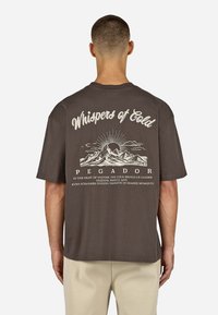 Hnedé tričko s krátkymi rukávmi, s grafickou potlačou hory a textom na chrbte. Bavlnený materiál, voľný strih, minimalistický dizajn.