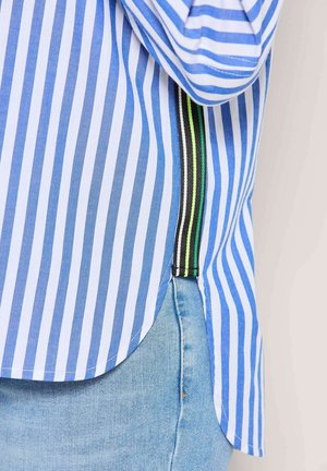 Blauw-wit gestreept shirt met een gebogen zoom, met een zwart-groen gestreepte accent aan de zijkant. Het materiaal lijkt licht en gestructureerd.