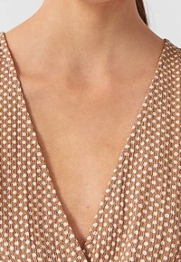 Gros plan sur une personne portant un vêtement à col en V texturé beige et blanc à pois, montrant le cou et le haut de la poitrine.