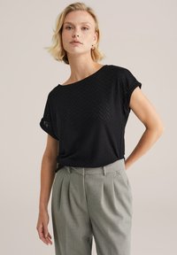 Černý vzorovaný krátký rukáv top s širokým výstřihem, kombinovaný s světle šedými kalhotami na míru. Texturovaná tkanina s herringbone vzorem.
