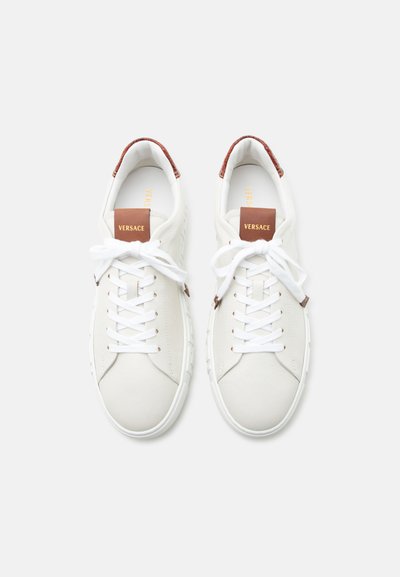 Paire de baskets en cuir blanc avec lacets blancs, languettes de talon marron, et étiquettes de languette marron embossées du mot « Versace » en lettres dorées.