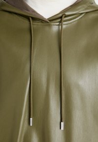 Haut à capuche en cuir vert olive avec un intérieur en tissu mélangé, cordons de serrage avec accents métalliques, et une texture lisse, légèrement brillante.