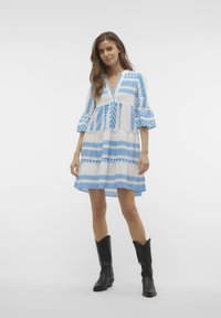 Robe blanche avec des motifs géométriques bleus, décolleté en V et manches volantes. Associée à des bottes de cow-boy noires. Tissu texturé.