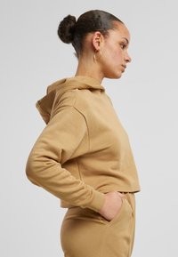 Sudadera con capucha recortada en tejido color tostado, con mangas largas y puños acanalados. Textura suave con un ajuste relajado, combinada con pantalones a juego.