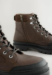Braune Lederstiefeletten mit strukturierter Oberfläche, ausgestattet mit einem seitlichen Reißverschluss, Metallbeschlägen und einer robusten Gummisohle mit Profil.