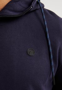 Sudadera con capucha de color Navy, hecha de una tela suave, que presenta un parche de logo negro. Los cordones de acento azul completan el diseño. Vista de cerca.
