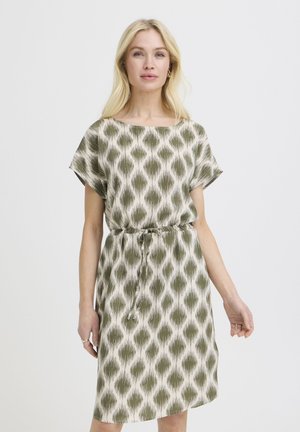 Blonde Frau in knielangem, weißem Kleid mit olivgrünen, abstrakten Rautenmuster und einem Bindegürtel, stehend vor einem schlichten Hintergrund.