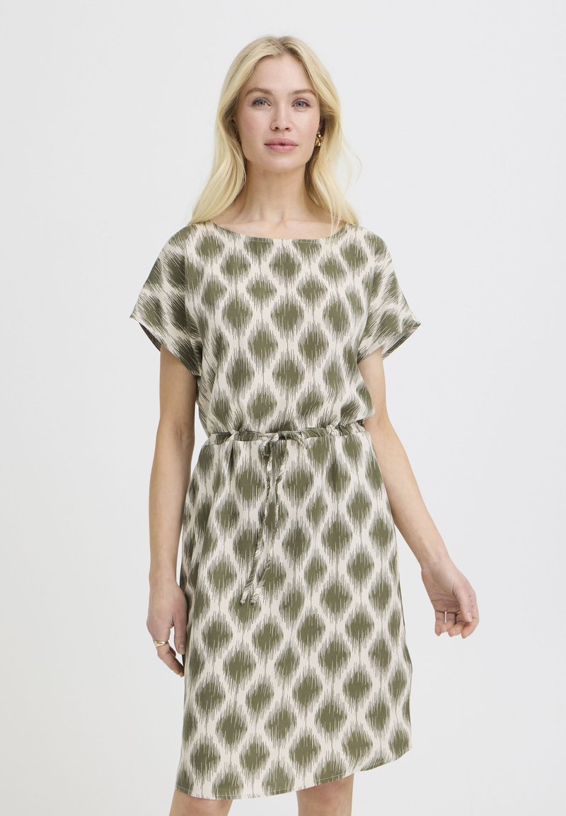 Blondynka w białej sukience do kolan z oliwkowo-zielonymi abstrakcyjnymi wzorami w kształcie diamentów i paskiem do wiązania, stojąca na jednolitym tle.