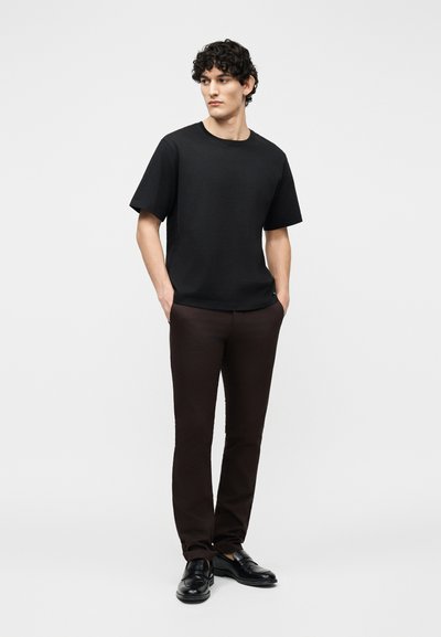 Jeune homme aux cheveux bouclés portant un t-shirt noir, un pantalon marron foncé et des chaussures en cuir noires, debout les mains dans les poches.