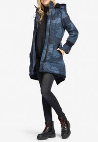 Kvinde iført en blå camouflage-quiltet vinterjakke med sorte leggings og sorte snørebodstøvler, stående med hænderne i lommen.