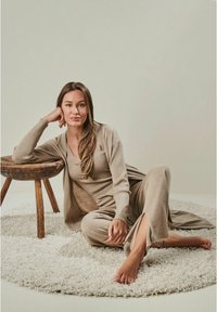 Beiges Strick-Loungewear-Set mit einem langen Cardigan und langen Hosen, die seitliche Schlitze aufweisen. Modell sitzt auf einem strukturierten weißen Teppich neben einem Holztisch.