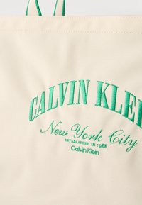 Μπεζ τσάντα χειρός Calvin Klein με πράσινο κεντητό κείμενο "Calvin Klein New York City Established in 1968" και ορατές πράσινες λαβές.
