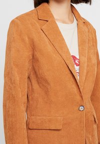 Benvävd kavaj i varm orange, med sned krage, en knappstängning och två sidofickor. Strukturerad yta med en mjuk känsla.