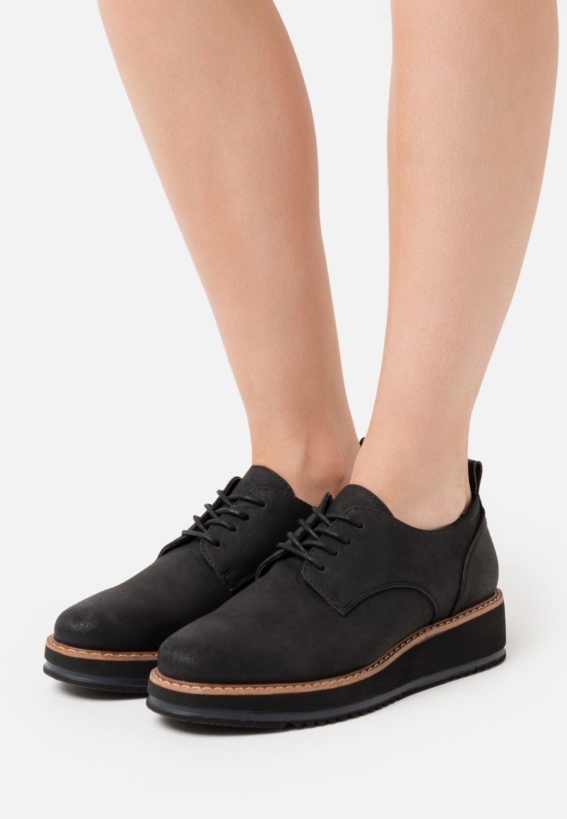 Jambes portant des chaussures à plateforme noires à lacets avec une bordure marron, présentées sur un fond clair uni.
