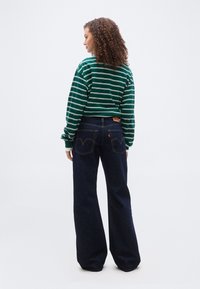 Γυναίκα με σγουρά μαλλιά, που φοράει μακρυμάνικο πουκάμισο με πράσινες και λευκές ρίγες και σκούρα τζιν παντελόνια με φαρδύ πόδι, στέκεται με την πλάτη γυρισμένη σε λευκό φόντο.